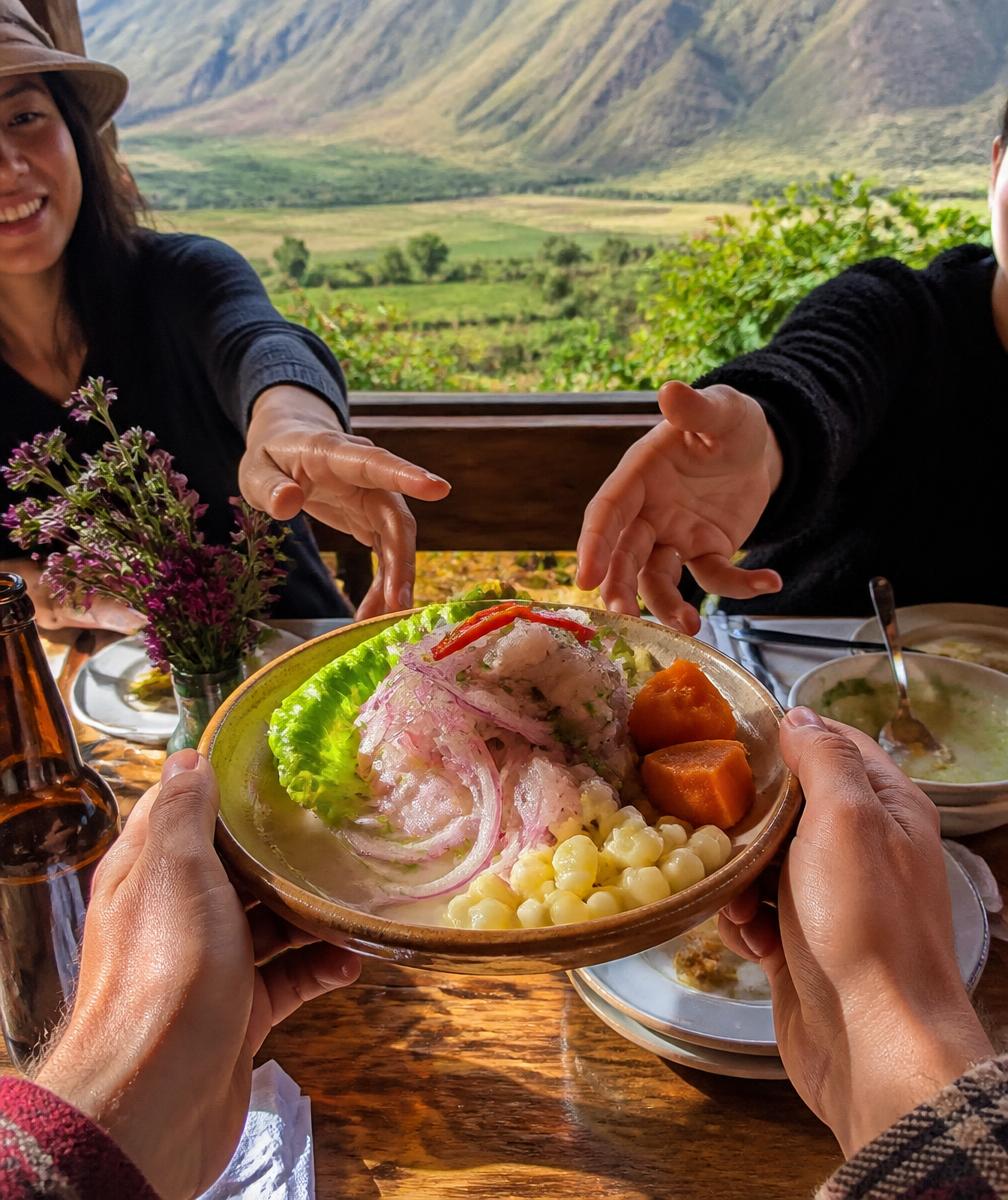 Manos compartiendo un plato de comida peruana en una mesa con vista al valle